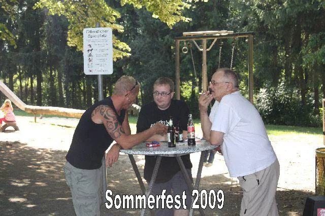 Drei Männer stehen um einen Stehtisch im Park, trinken und sprechen. Es ist Sommer 2009, Sommerfest, im Hintergrund Spielplatz.