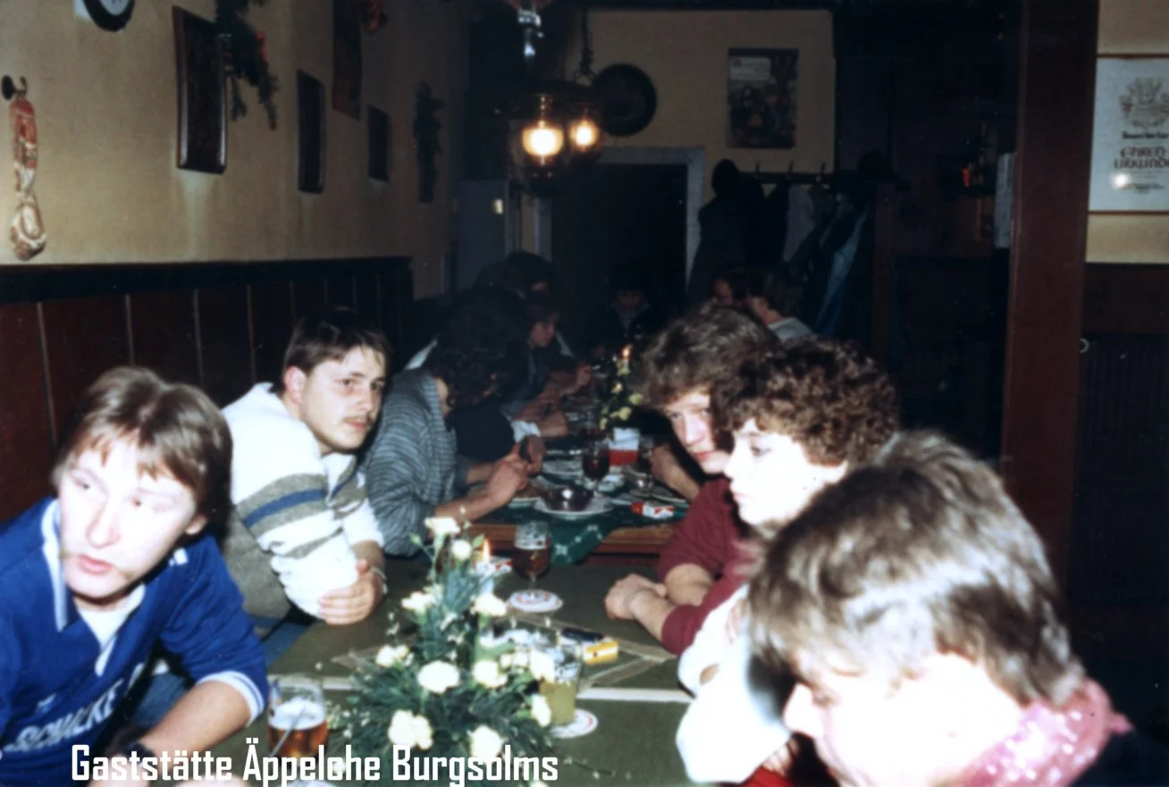 Mehrere Personen sitzen an einem langen Tisch in einem rustikalen Restaurant. Der Tisch ist mit einer grünen Tischdecke und einem Blumenarrangement in der Mitte gedeckt. Im Hintergrund hängen Wände mit Dekorationen und es gibt eine Beleuchtung an der