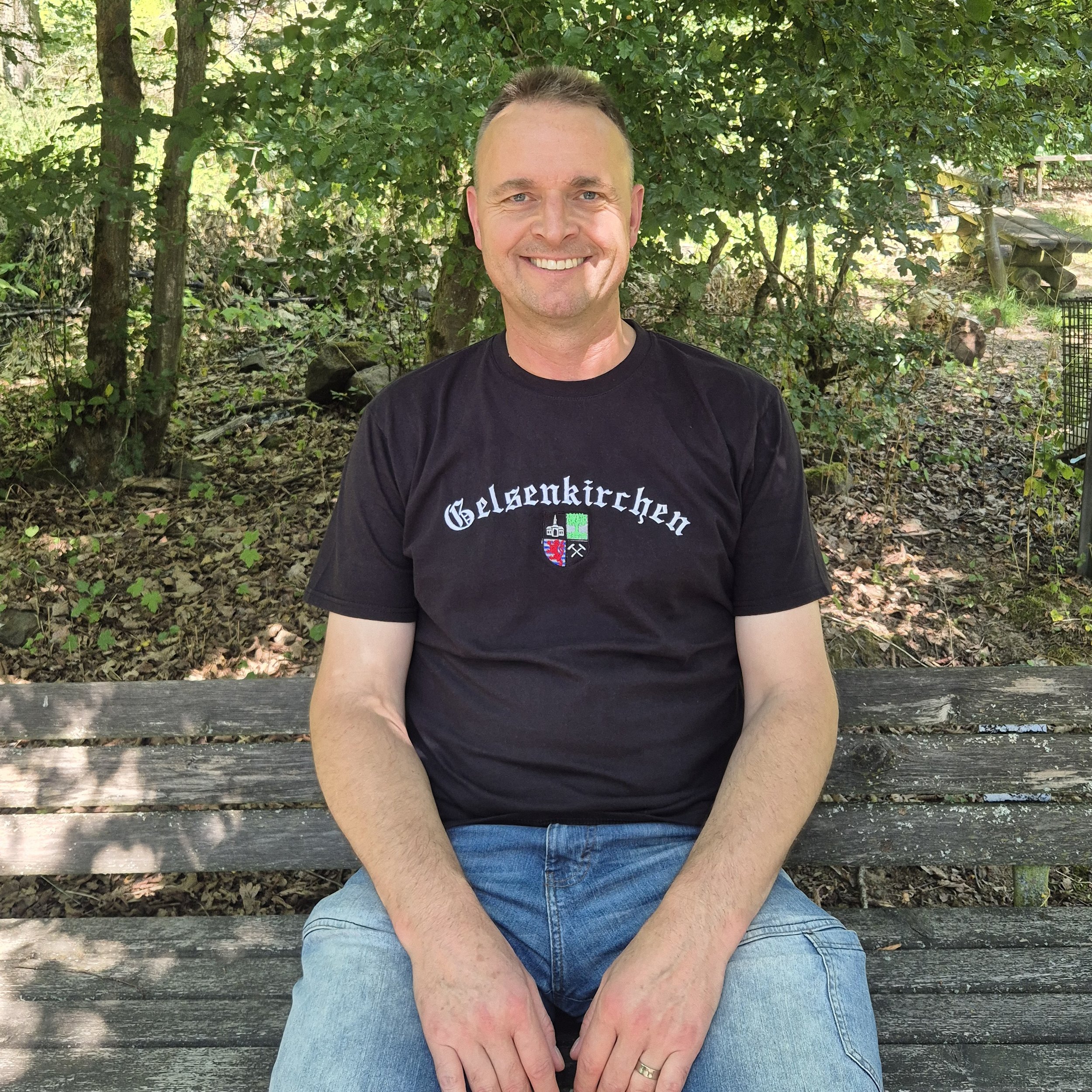 Ein Mann sitzt auf einer Bank im Freien, umgeben von Bäumen und Laub, trägt ein schwarzes T-Shirt und blaue Jeans, lächelt.