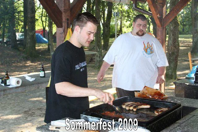 Zwei Männer grillen Würstchen und Fleisch auf einem offenen Grill bei einem Sommerfest im Jahr 2009 im Freien, umgeben von Bäumen.