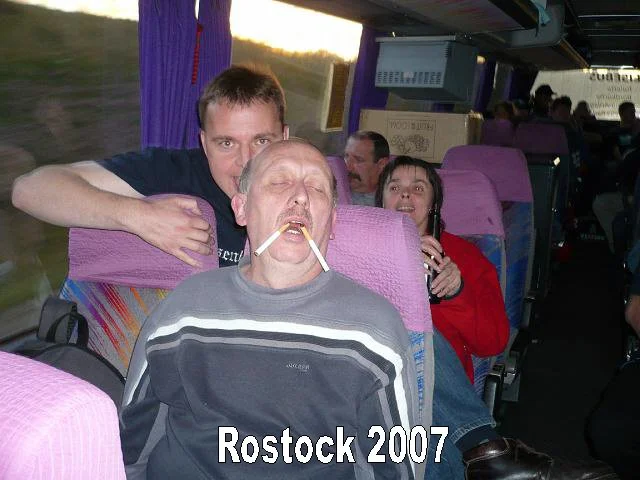 Gruppe von Menschen in einem Bus, eines hält eine Zigarette im Mund, im Hintergrund lachen andere.