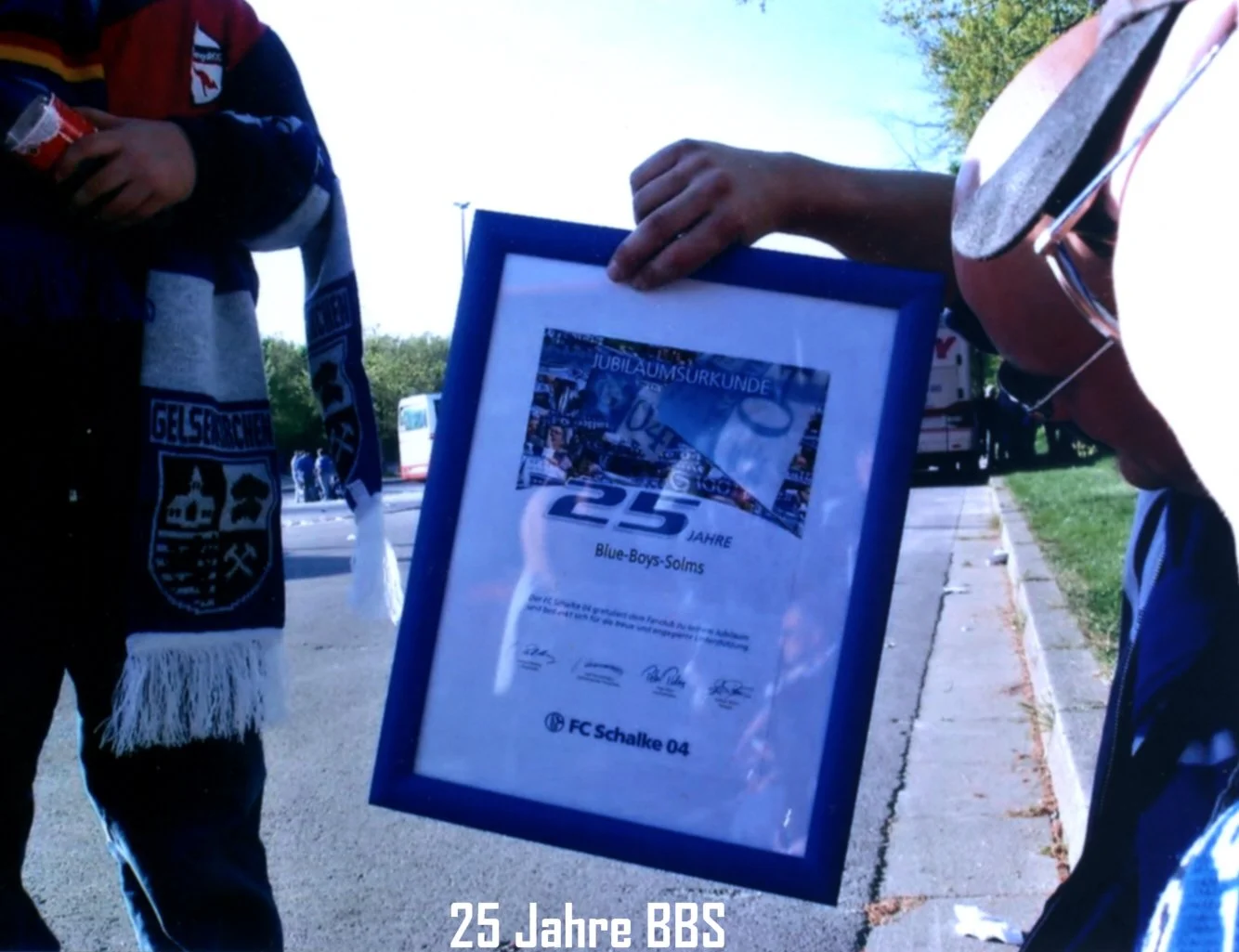 Zwei Personen halten ein gerahmtes Dokument mit der Aufschrift "25 Jahre Blue-Boys-Solms" bei einer Jubiläumsfeier. Im Hintergrund sind Bäume, ein Bus und eine Straße zu erkennen.