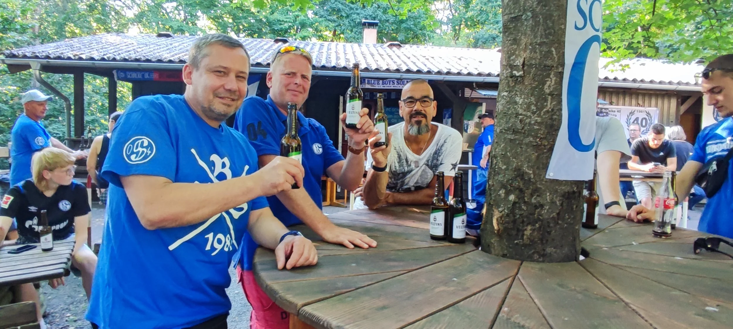 Menschen in blauen Trikots feiern und trinken Bier an einem Holzpult im Freien, umgeben von Bäumen und anderen Personen, mit einem Baum in der Mitte des Pults.