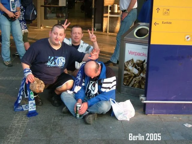 Drei Männer sitzen auf dem Boden vor einem Bahnhof, einer hält eine Flasche, die anderen machen Peace-Zeichen. Es ist Berlin 2005.