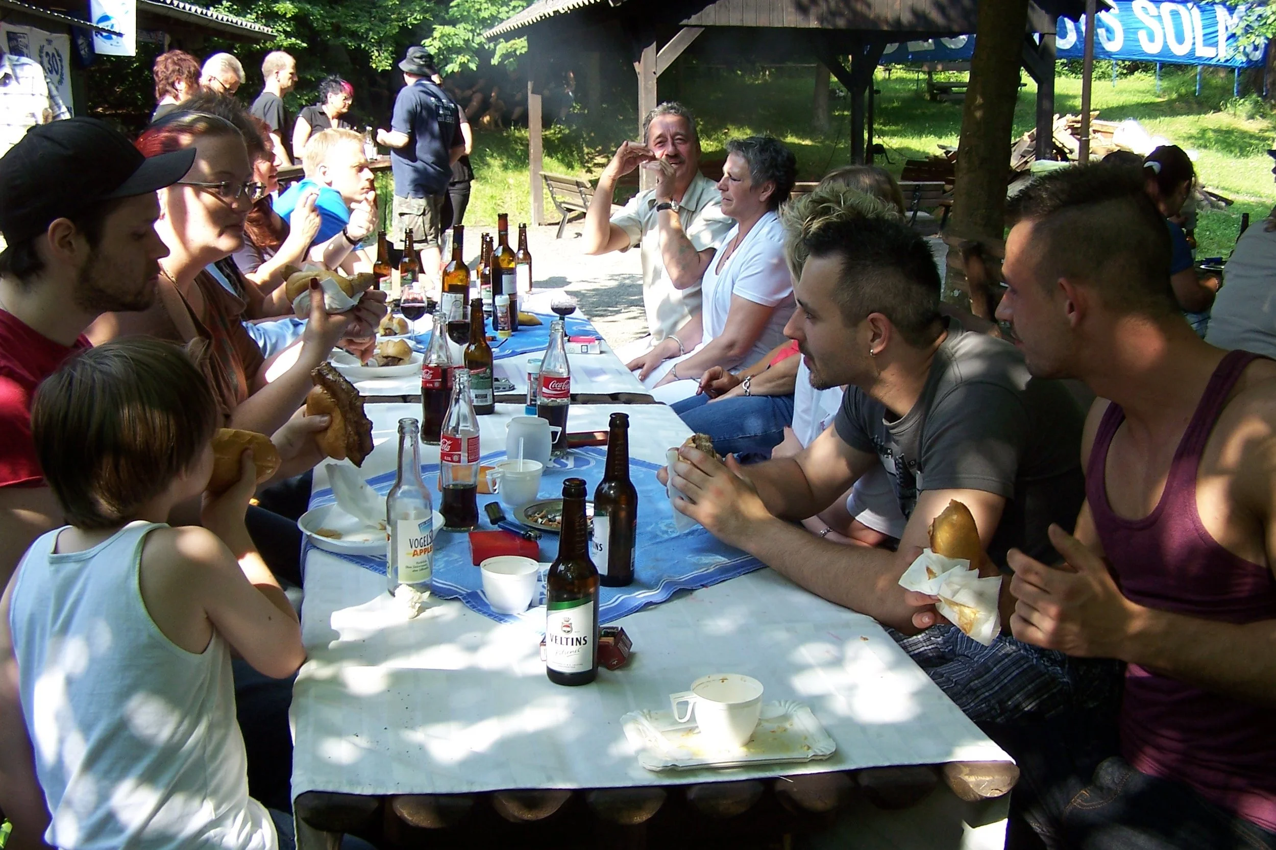 Menschen sitzen an einem langen Tisch im Freien bei einer geselligen Zusammenkunft, essen, trinken und unterhalten sich unter Schatten spendenden Bäumen.