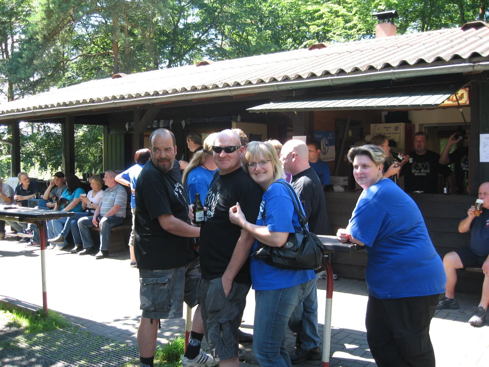 Gruppen von Menschen, die draußen an einem Barbereich zusammenstehen und trinken, bei einem Treffen oder Festival an sonnigem Tag, mit Wald im Hintergrund.