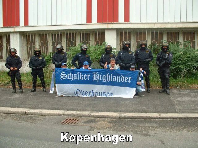 Gruppen von Männern in dunklen Polizeiuniformen mit Helmen stehen hinter einer Gruppe von drei Personen, die ein blau-weißes Banner halten, auf dem 'Schalker Highlander Oberhausen' steht, vor einer Mauer mit Pflanzen. Das Bild ist im Stadtgebiet Kope