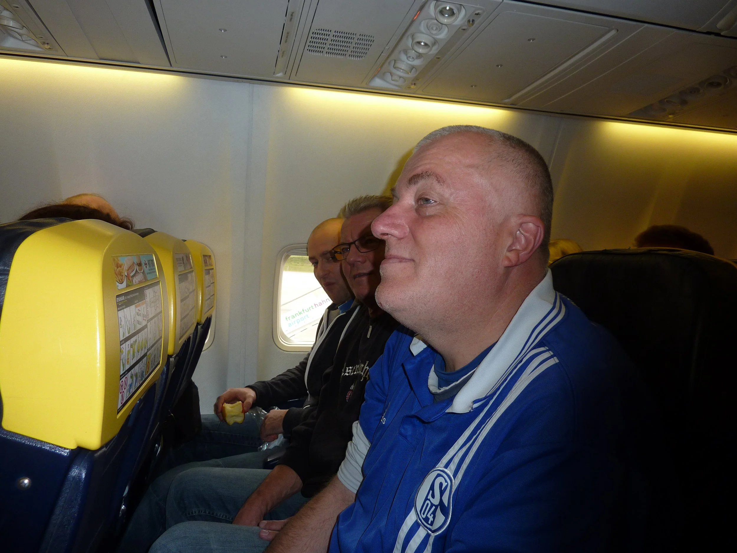 Menschen sitzen in einem Flugzeug. Eine Person im Vordergrund trägt ein blauen Sport-Trikot mit einem Logo. Die Sitze sind gelb und oberhalb gibt es eine Notausgangsbroschüre. Die Passagiere wirken entspannt.