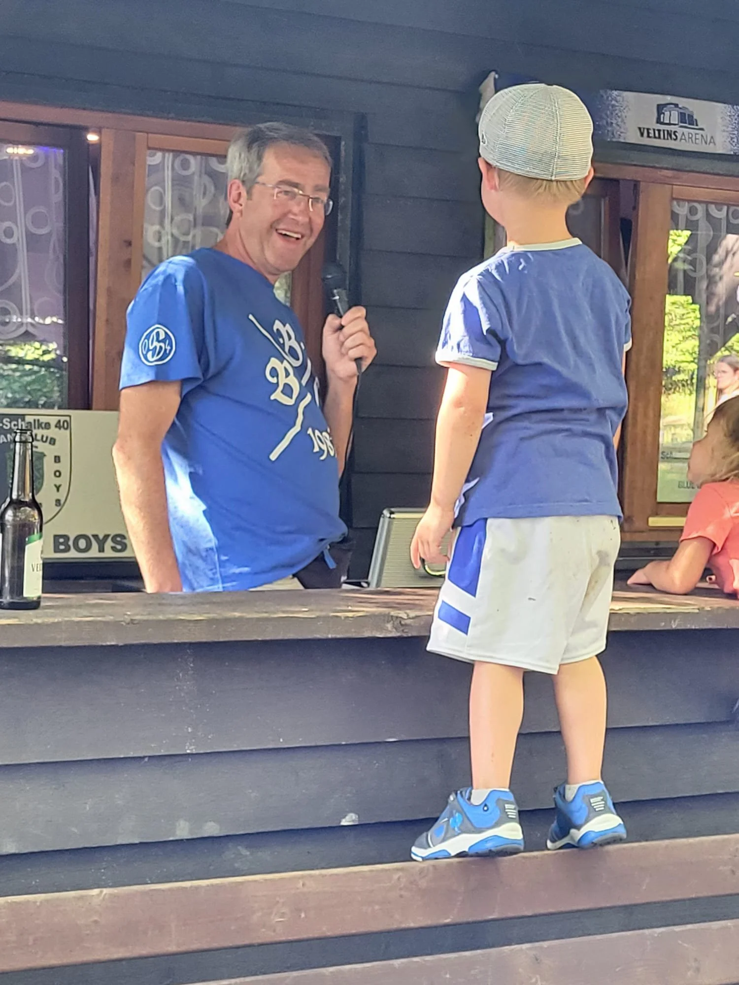 Ein Mann in blauer Trikot führt ein Gespräch mit einem jungen Jungen in Sportkleidung und Turnschuhen an einem Stand bei einem Outdoor-Turnier.