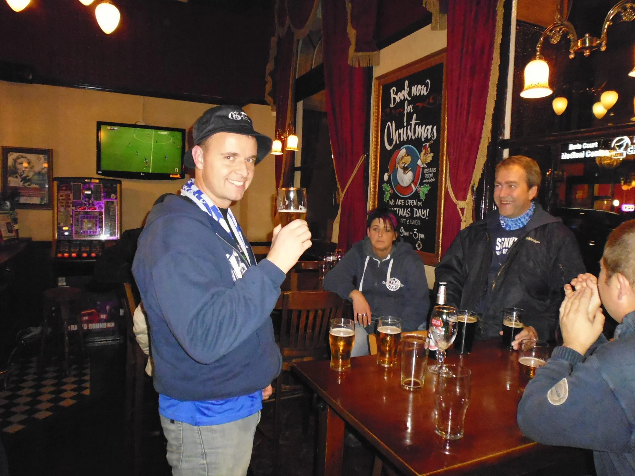 Menschen in einer Bar trinken Bier und unterhalten sich. Es gibt eine Wand mit einem Weihnachtsplakat und einem flachen Bildschirm, der ein Fußballspiel zeigt.