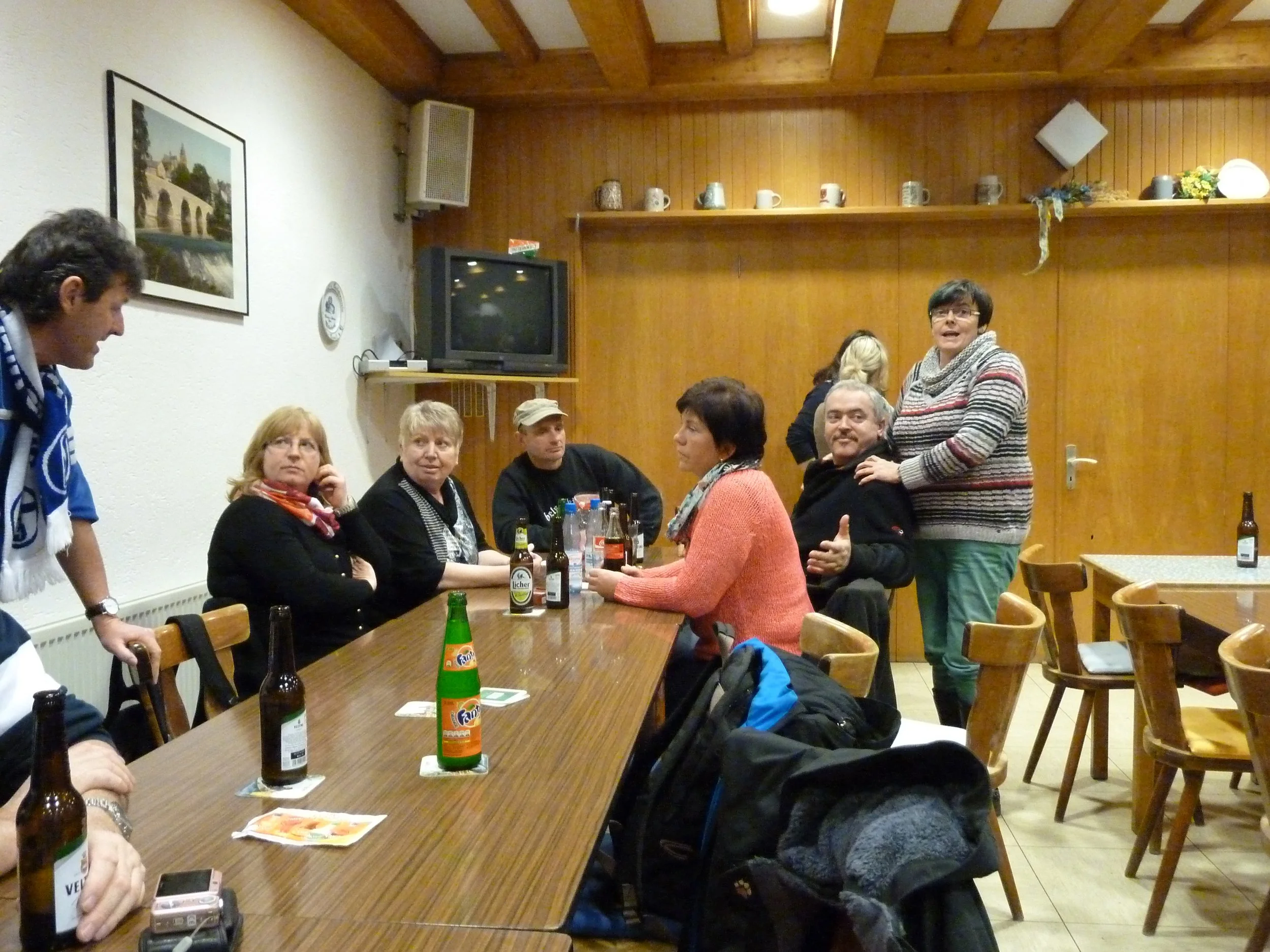 Mehrere Menschen sitzen um einen langen Tisch in einem Raum mit Holzdecke und Wänden, trinken Getränke und unterhalten sich, während einige andere stehen.