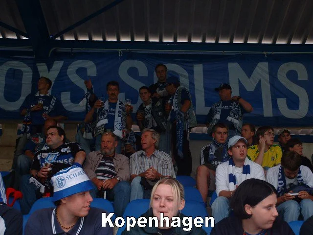 Fußballfans in einem Stadion, einige tragen Schals und Trikots des FC Schalke 04, im Hintergrund eine Bande mit der Aufschrift 'OS S OLM S', darunter der Titel 'Kopenhagen'.