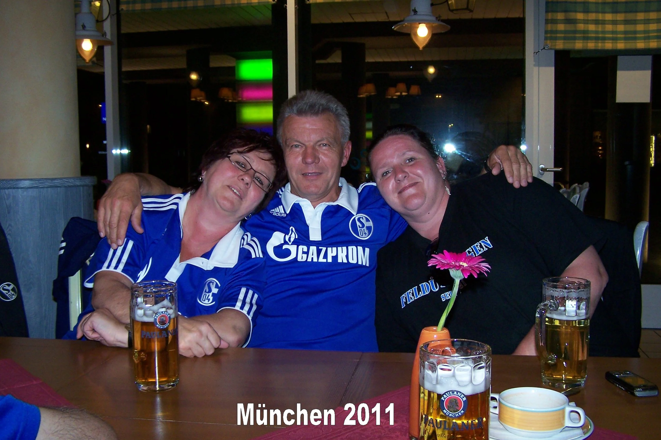 Drei Menschen, zwei Frauen und ein Mann, sitzen in einer Kneipe oder einem Restaurant. Sie tragen blaue Trikots mit dem Logo von Schalke 04. Sie sind fröhlich und haben die Arme umeinander gelegt. Auf dem Tisch vor ihnen stehen Biergläser, einige dav