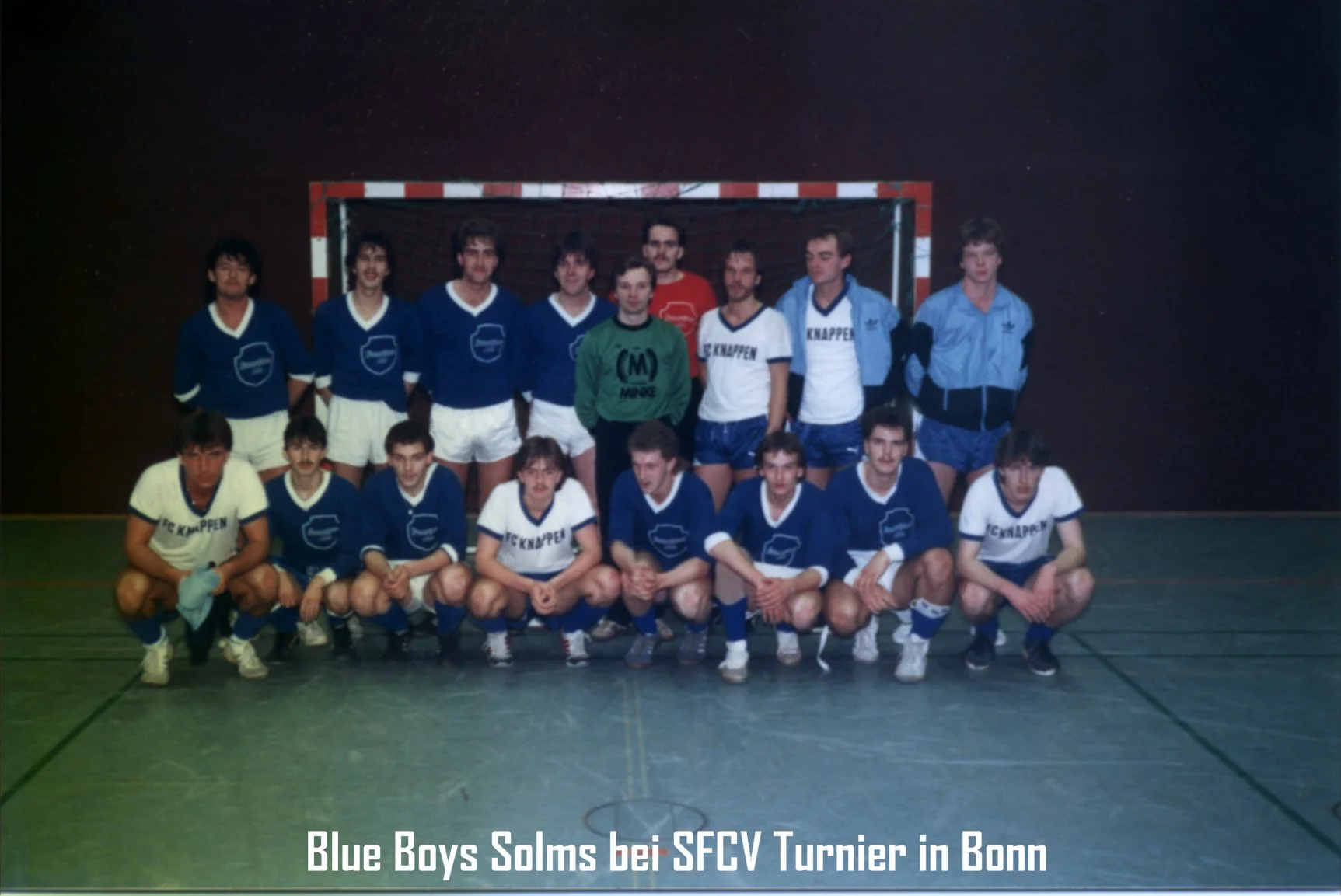 Eine Gruppe von jungen Männern in blauen und weißen Fußballtrikots, Fußballspieler, posieren vor einem Tor in einer Turnhalle, Hintergrund ist dunkel, auf dem Boden Linien für den Hallenfußball, unten Beschriftung: "Blue Boys Solms bei AFCV Turnier i