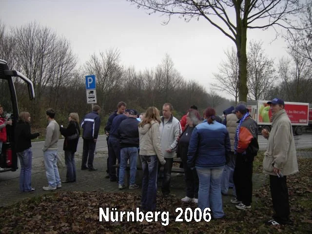 Gruppe von Menschen steht an einer Landstraße in Nürnberg im Jahr 2006, einige warten, andere sprechen miteinander, umgeben von Bäumen und einem LKW im Hintergrund.