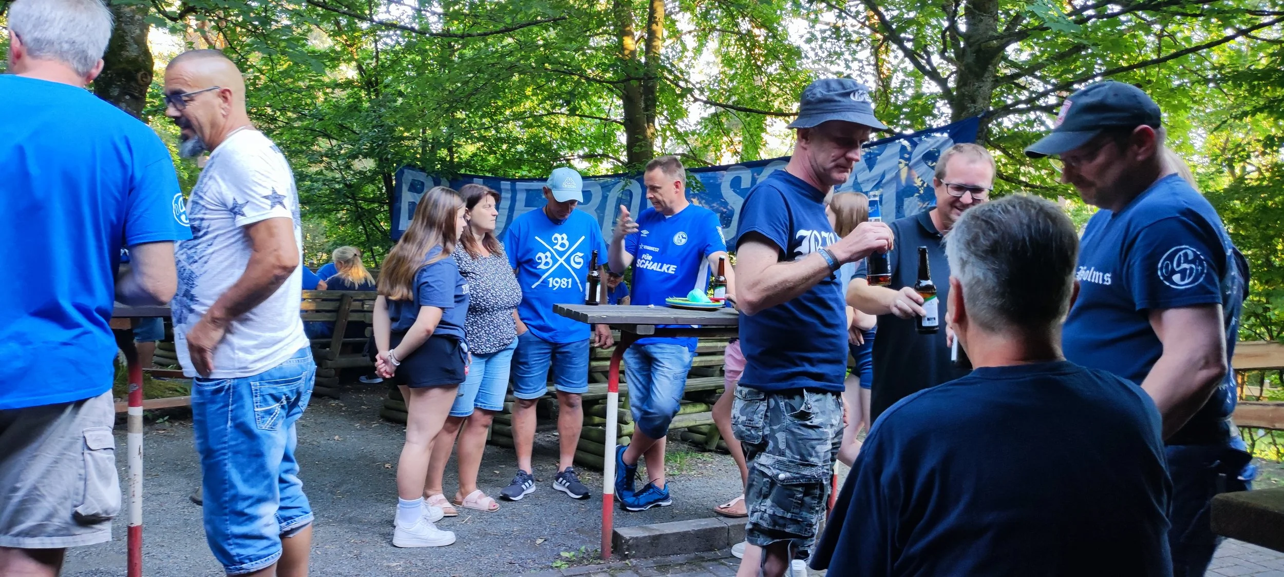 Gruppen von Menschen, die im Freien in einem grünen, bewaldeten Bereich zusammenstehen, mit einigen, die Bier trinken und sich unterhalten.