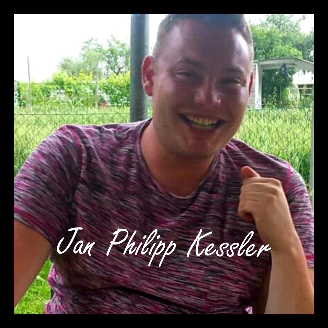 Ein lachender Mann in einem roten gestreiften T-Shirt, im Hintergrund ein grüner Garten mit Bäumen und einer Zäunung.