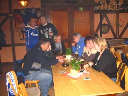 Mehrere Menschen sitzen und stehen in einer Bar, einige schauen auf ihre Handys, andere unterhalten sich, es gibt Getränke auf dem Tisch.