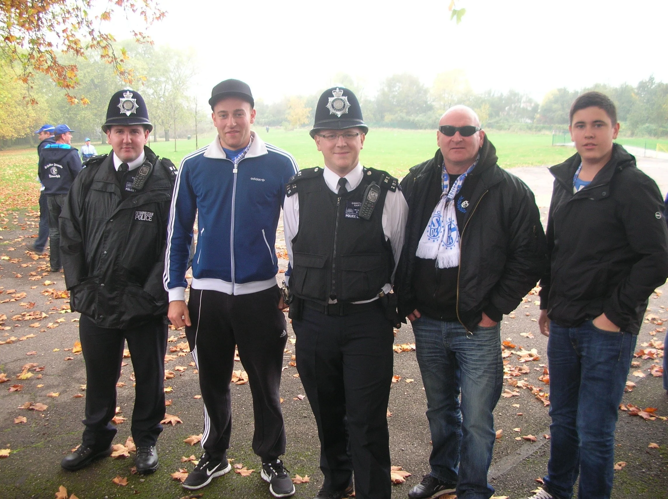 Fünf Männer stehen zusammen im Park, einige tragen Polizei-Uniformen mit Mützen. Es befindet sich Herbstlaub auf dem Boden, im Hintergrund sind Bäume und mehr Menschen, einige in blauen Jacken.