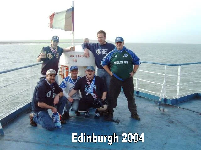Eine Gruppe von sieben Menschen steht auf einem Schiff, das im Meer fährt. Sie tragen Sportkleidung und Hüte, ein Mann hält einen Regenschirm mit dem Logo Veltins. Im Hintergrund ist eine Flagge und ein Mast. Der Text unten lautet 'Edinburgh 2004'.