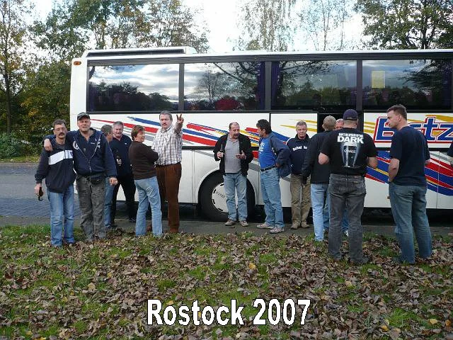 Gruppe von Männern steht vor einem Bus auf einer Wiese mit Herbstlaub, im Hintergrund Bäume und Himmel übersät mit Wolken. Der Text "Rostock 2007" ist unten im Bild.