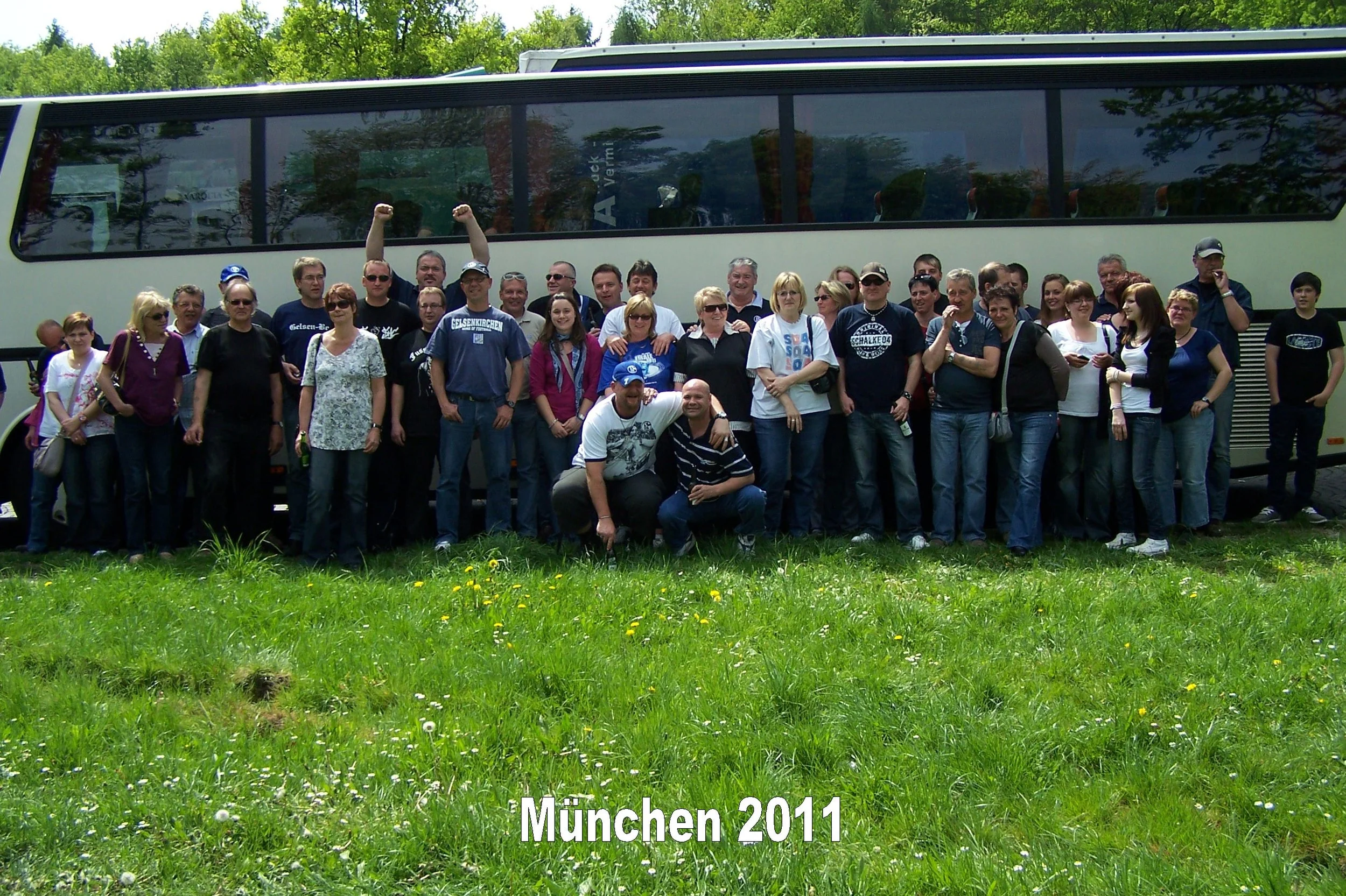 Eine Gruppe von Menschen vor einem Bus, die auf einem grünen Rasen mit Blumen stehen, mit Bäumen im Hintergrund. Der Text unten liest 'München 2011'.