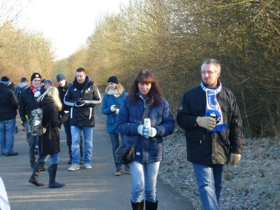 Gruppe von Menschen in Winterkleidung, die draußen an einer Straße stehen, einige halten Getränke und schauen in verschiedene Richtungen, im Hintergrund Bäume.