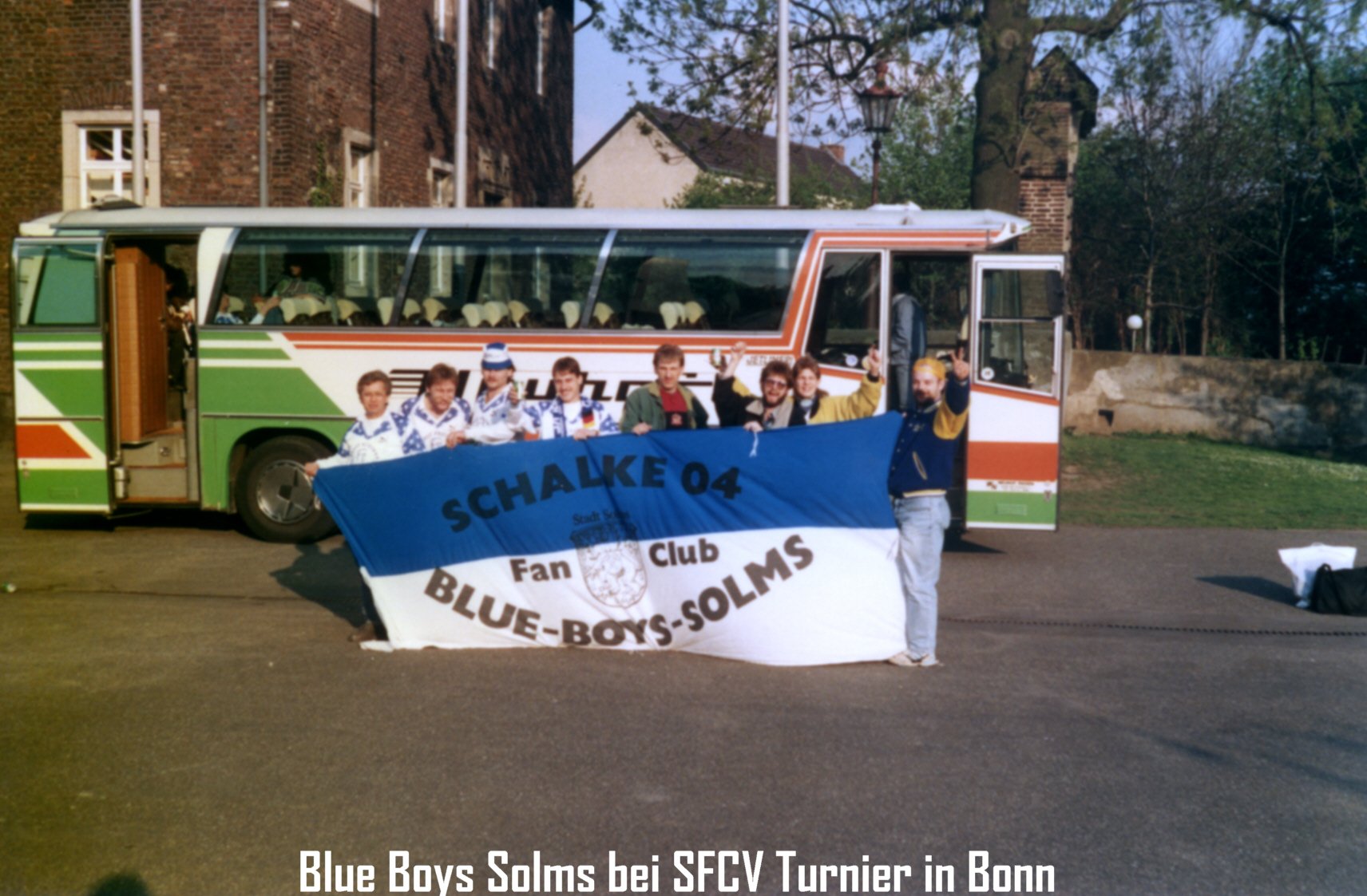 Gruppe von Menschen vor einem Bus, die einen blauen und weißen Fan-Club-Teppich halten, mit einem Banner, auf dem 'SCHALKE 04 BLUE BOYS SOLMS' steht, in Bonn.