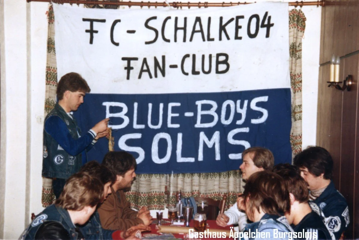 Ein Gruppenbild von mehreren jungen Menschen, die um einen Tisch sitzen, mit einer großen Fahne im Hintergrund, auf der steht 'FC-Schalker04 Fan-Club' und 'Blue-Boys Solms'.
