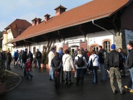 Menschen stehen in einer Warteschlange vor einer historischen Bahnhofshalle mit roten Ziegeldächern in Deutschland.