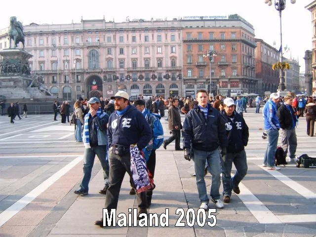 Menschen stehen und laufen auf einem öffentlichen Platz in Mailand im Jahr 2005, mit historischen Gebäuden und einer Statue im Hintergrund.