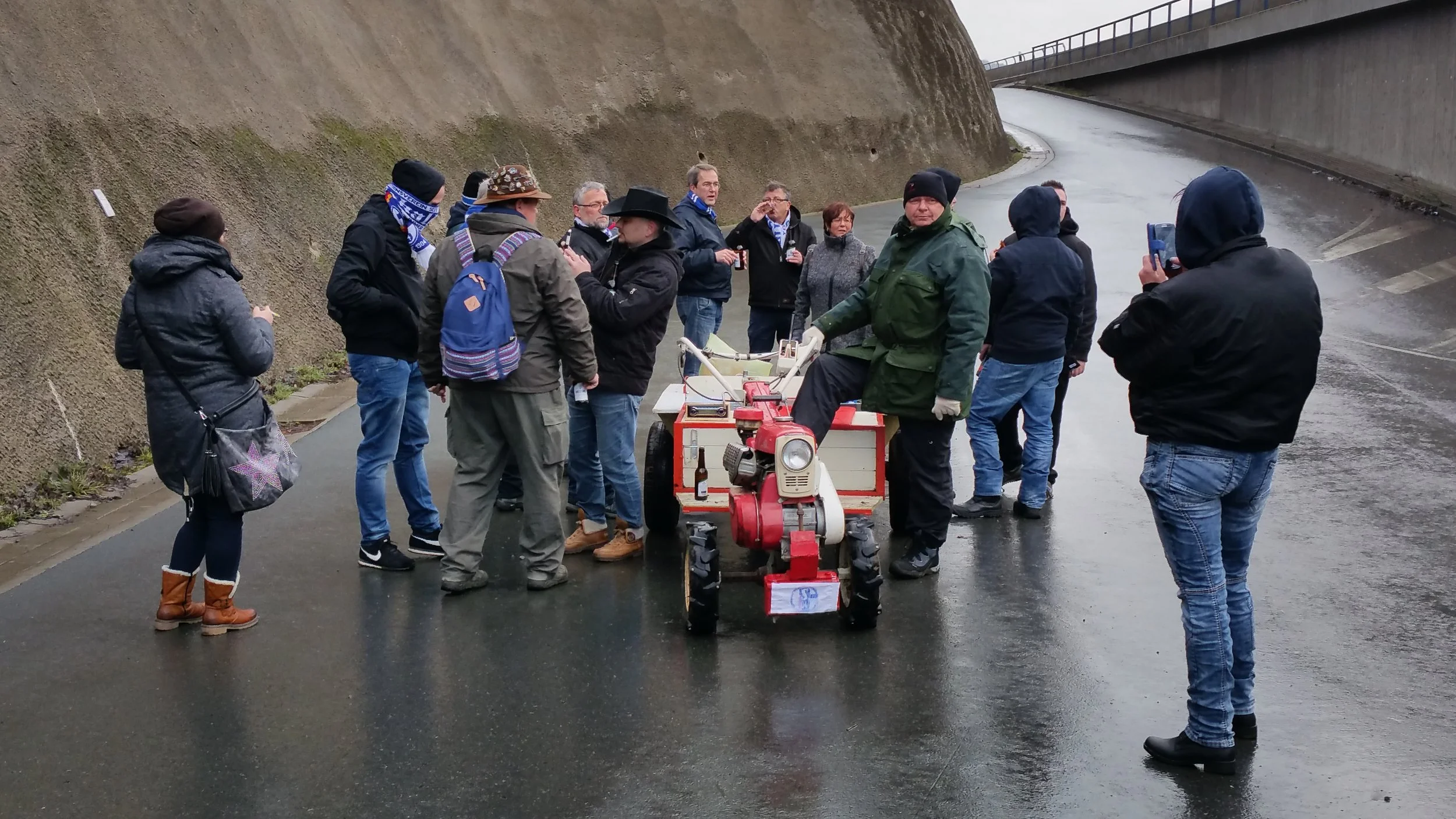 Eine Gruppe von Menschen steht auf einer nassen Straße neben einem Wasserkanal, einige tragen warme Kleidung und schauen auf einen kleinen motorisierten Anhänger.
