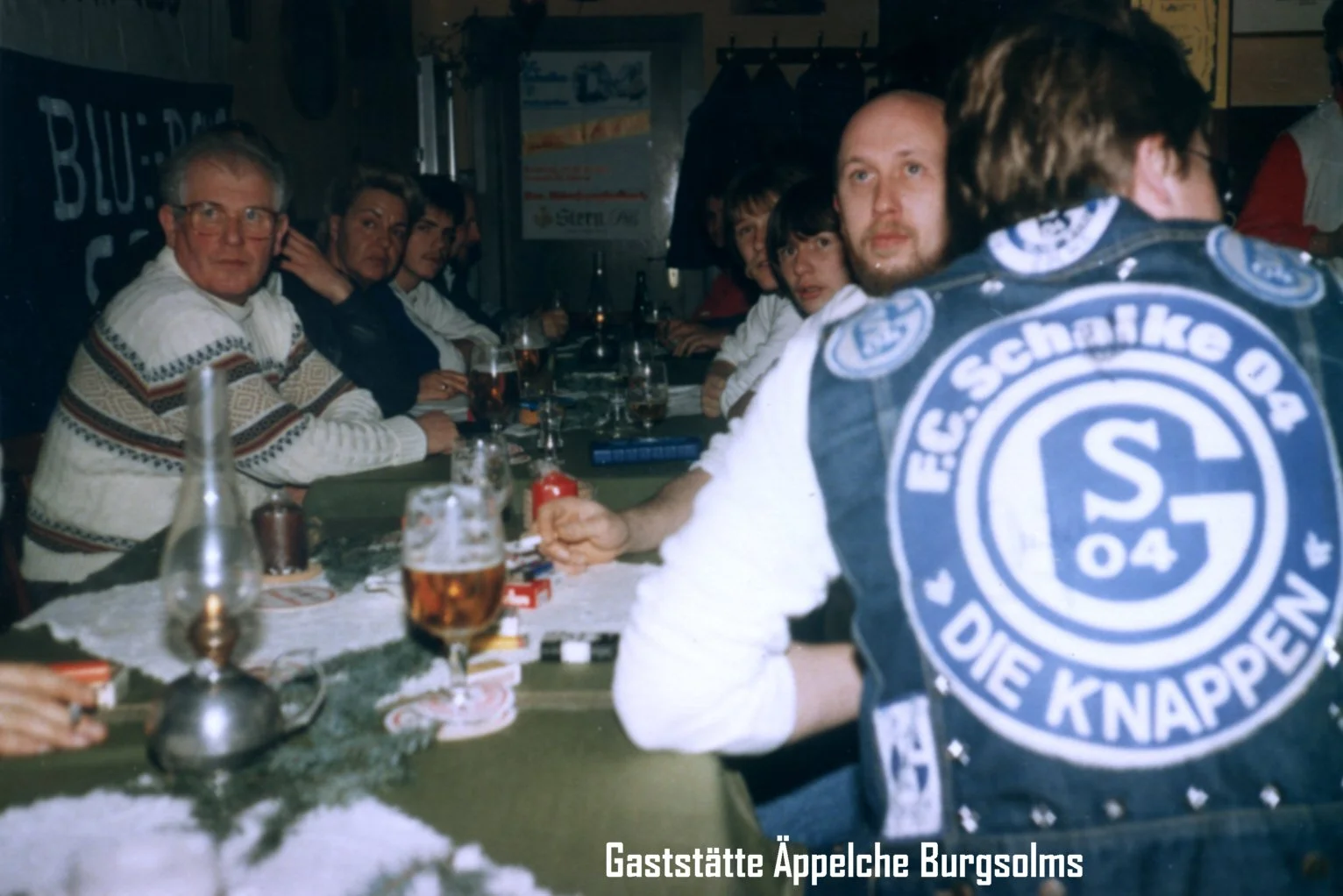 Menschen sitzen an einem Tisch in einer Gaststätte, einige tragen Bayern-Jacken, Getränke und Zigaretten sind auf dem Tisch. Der Ort heißt "Gaststätte Äppelche Burgsolms".