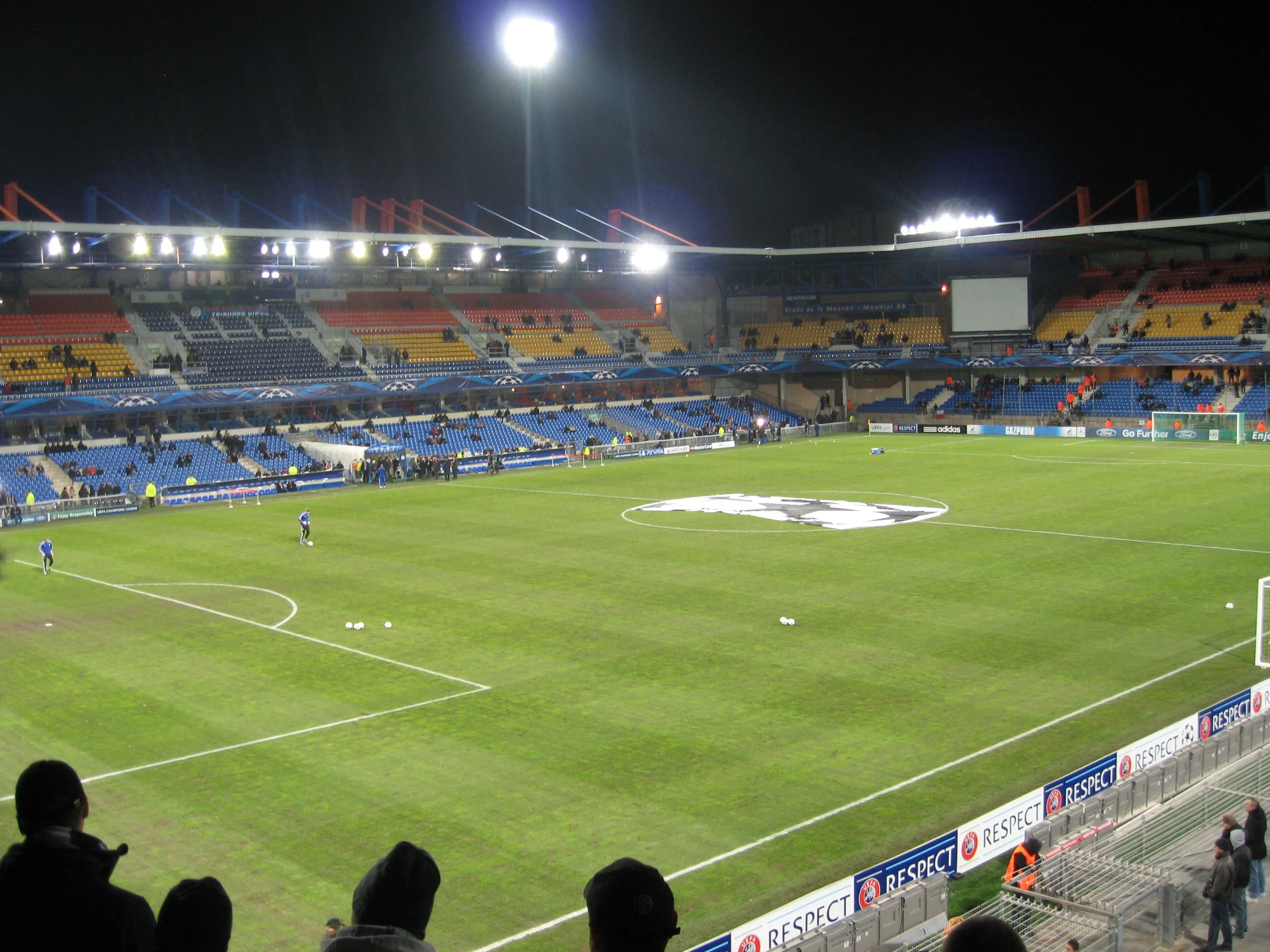 Ein Fußballstadion bei Nacht mit leuchtenden Flutlichtern, leeren Sitzplätzen und einem gepflegten Rasen, wo Spieler für das Spielvorbereitungen treffen.