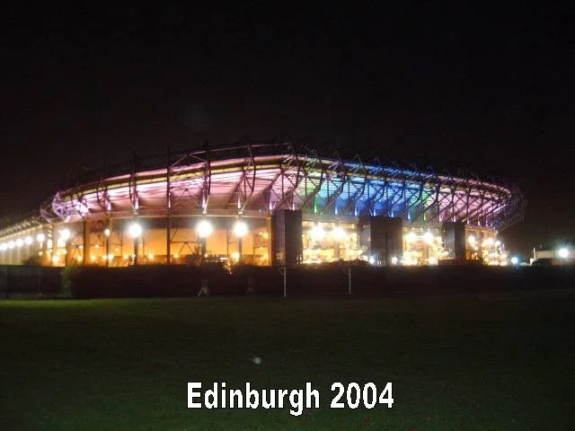 Ein beleuchteter Sportstadion bei Nacht mit bunten Lichtern, im Vordergrund eine Rasenfläche, darunter Text 'Edinburgh 2004'.