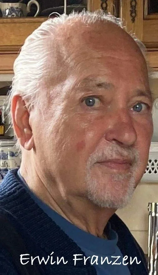 Nahaufnahme eines älteren Mannes mit weißen Haaren, blauen Augen und einem leichten Lächeln, im Hintergrund eine Küchenszene mit Schränken und Geschirr. Der Name "Erwin Franzen" ist unten im Bild geschrieben.