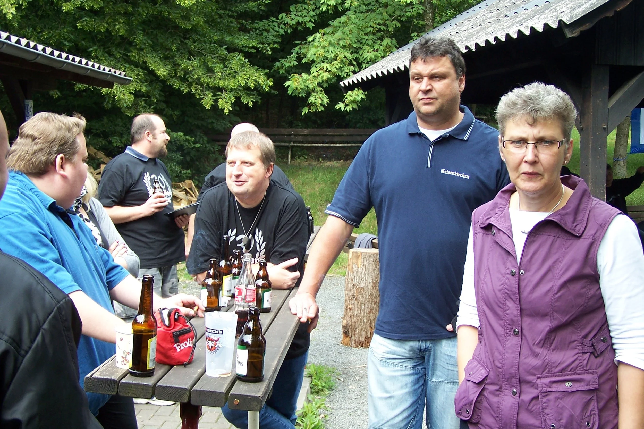 Gruppe von Menschen bei einem Treffen im Freien, einige sitzen an einem Tisch mit Getränken, andere stehen und unterhalten sich, im Hintergrund Bäume und ein Holzgebäude.