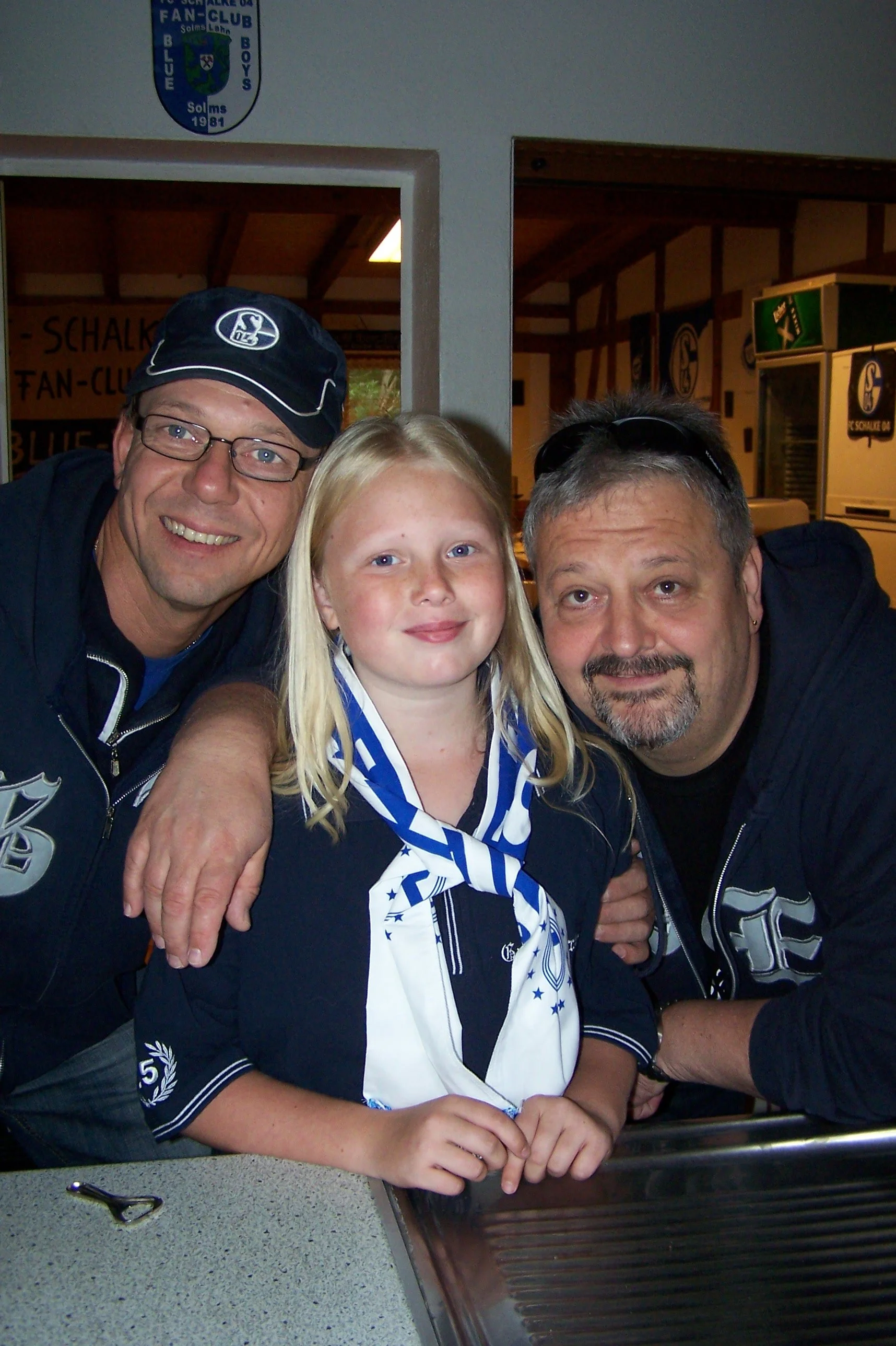 Zwei Männer und ein Mädchen, alle tragen Fan-Club Trikots, posieren für ein Foto. Im Hintergrund sind Fan-Club-Logos und eine Getränkeautomat. Es scheint ein Fan-Club-Treffen zu sein.