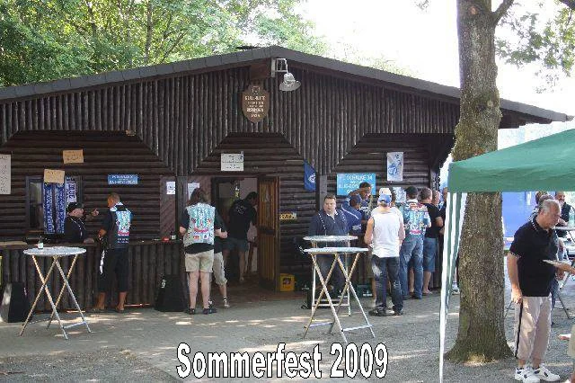 Menschen vor einem Holzgebäude beim Sommerfest 2009, einige stehen in einer Schlange, andere sitzen an Stehtischen, es gibt einen grünen Pavillon im Vordergrund