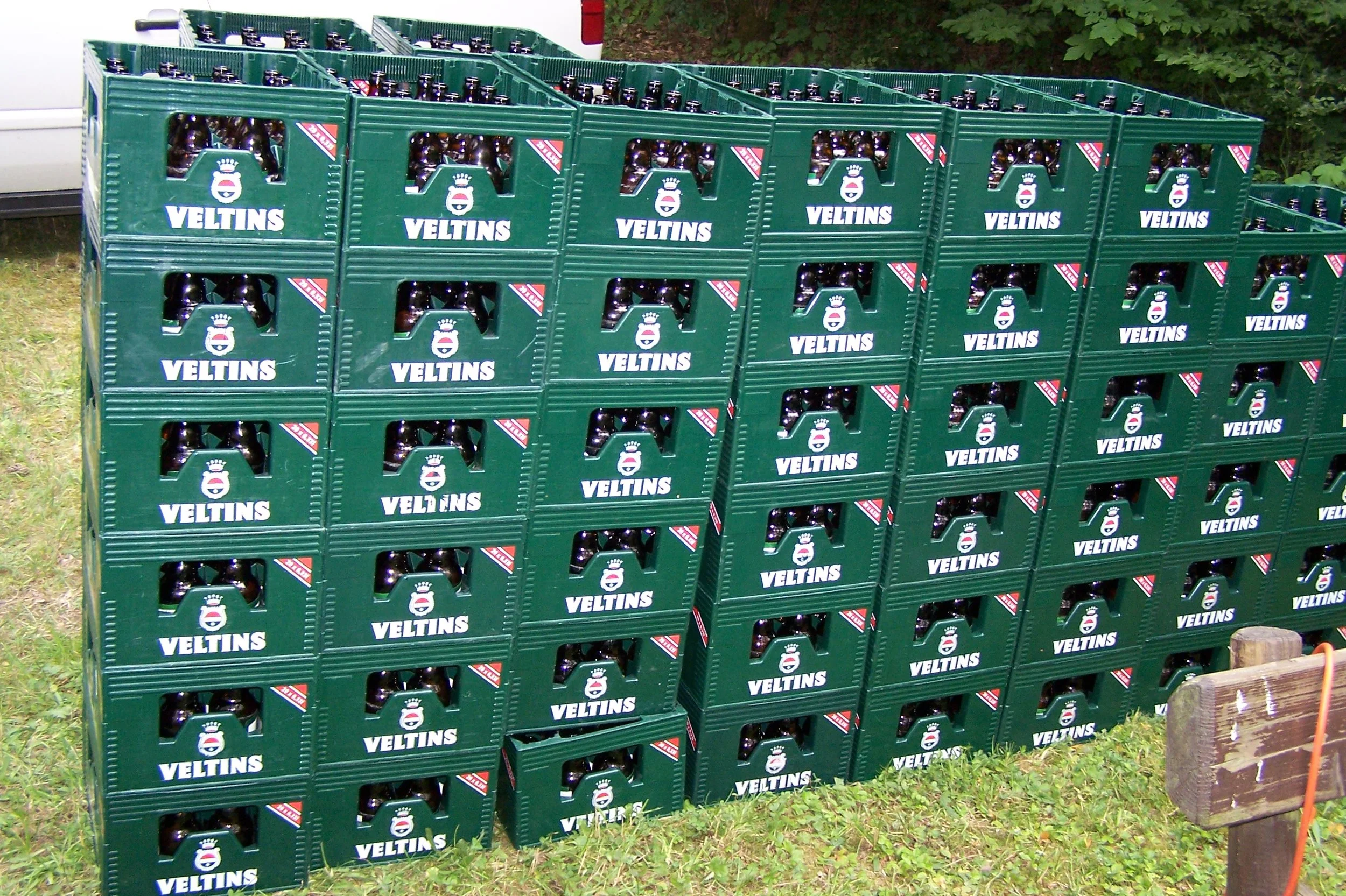 Mehrere grüne Kisten mit der Bezeichnung "Veltins", vollgefüllt mit Flaschen, stehen auf Gras neben einem Holzpfosten. Es handelt sich um eine Bierlagerung.