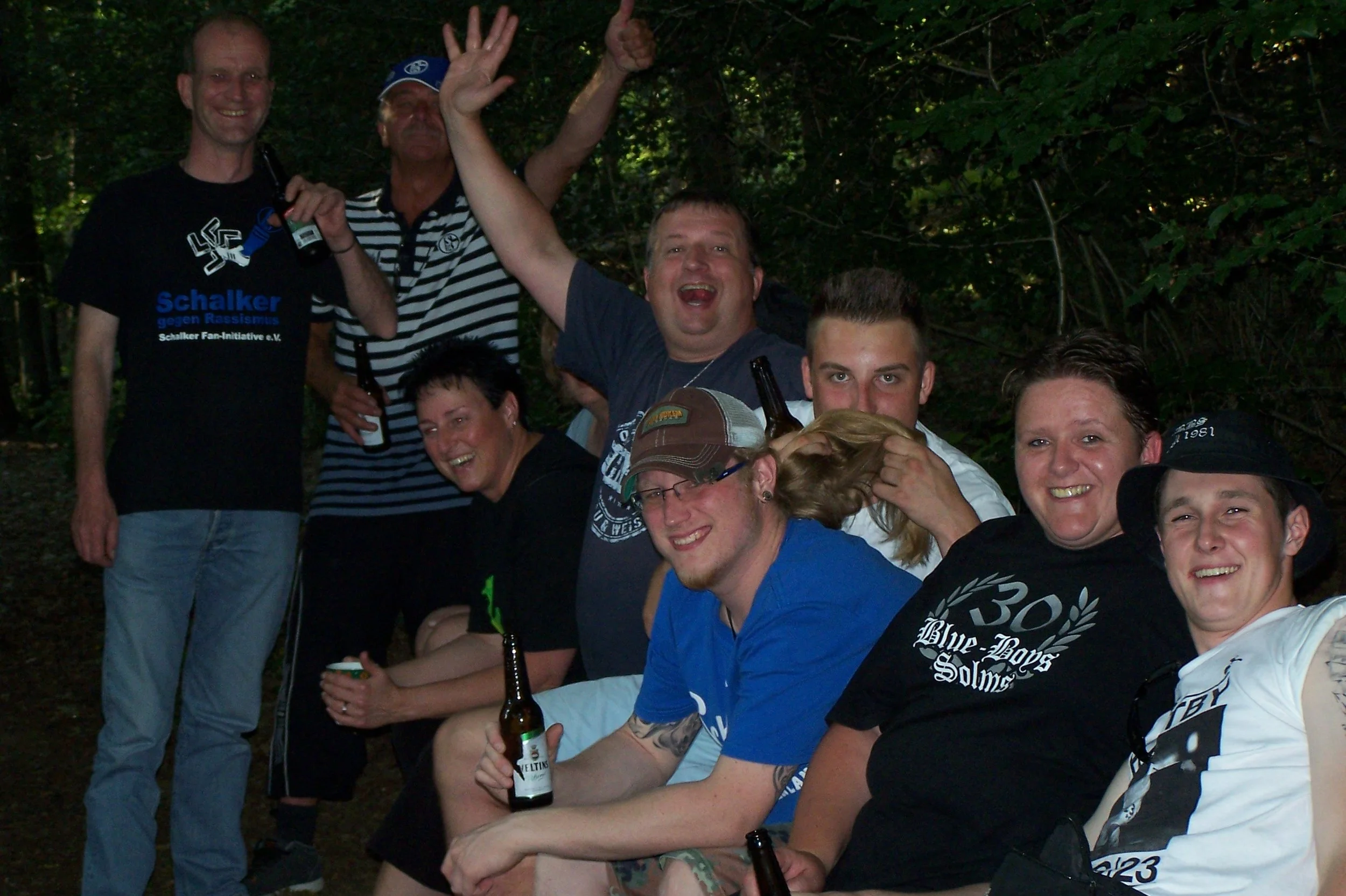 Gruppe von fröhlichen Menschen, die im Freien unterwegs sind, einige halten Getränke in der Hand, im Hintergrund sind Bäume zu sehen.