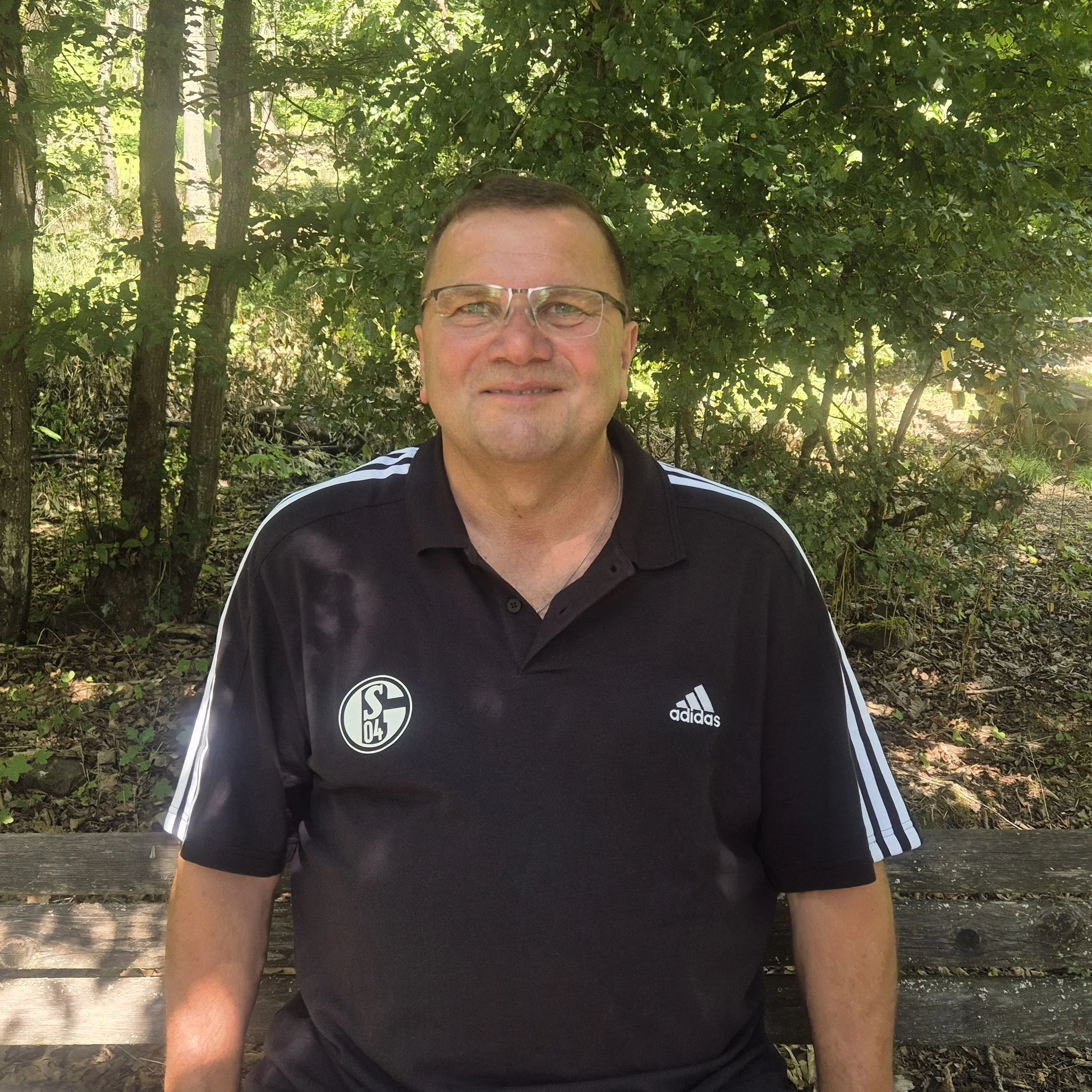 Nahaufnahme eines Mannes mit Brille, schwarzem Adidas-Shirt mit Schalke 04 Logo, sitzend an einem Baum im Grünen, im Freien an einem sonnigen Tag.