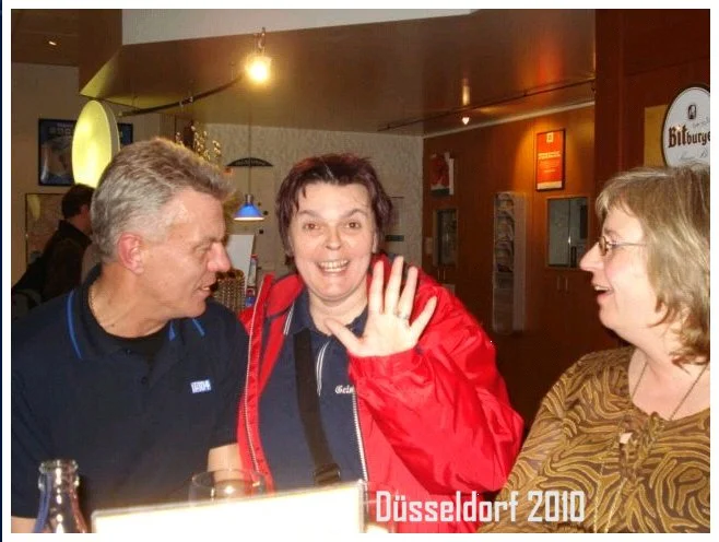 Drei Personen sitzen an einem Tisch in einer Bar oder Kneipe, lachen und unterhalten sich. Der Mittelperson winkt in die Kamera. Im Hintergrund sind Lampen und Wanddekorationen zu sehen. Text im Bild: "Düsseldorf 2010".