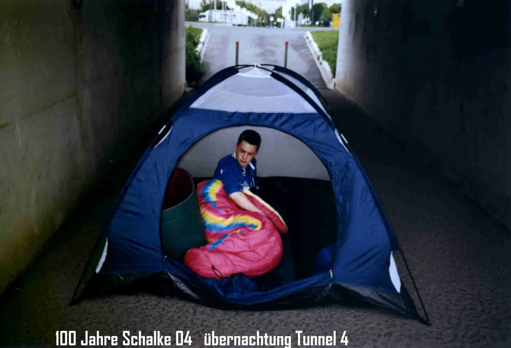 Ein Junge sitzt in einem Tunnel in einem Zelt und wickelt sich in eine bunte Decke ein, nach oben geöffnet, Helden/Bieracke im Hintergrund.