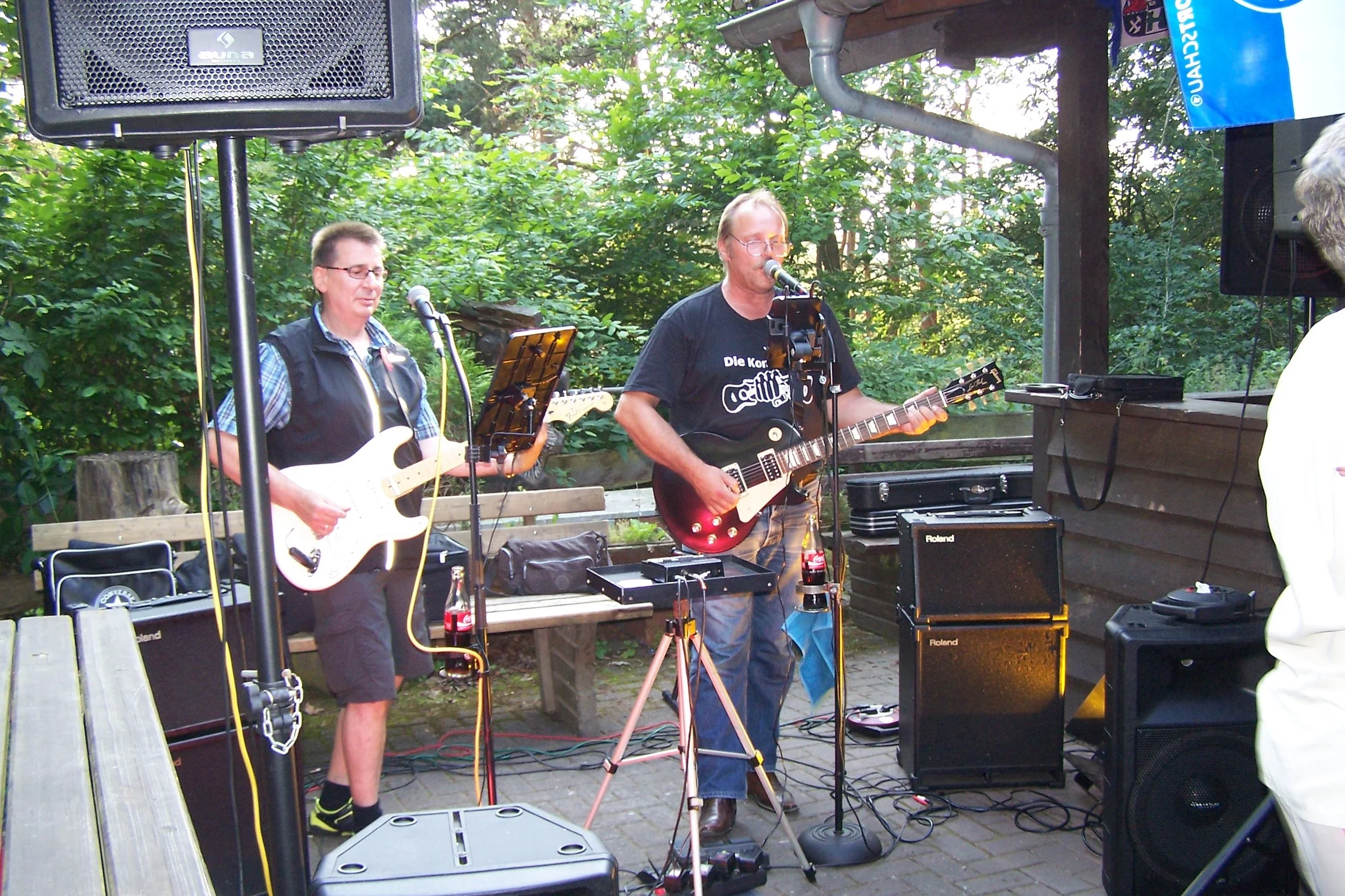 Zwei Musiker spielen Gitarre auf einer Bühne im Freien, umgeben von Bäumen, mit Mikrofonen, Verstärkern und Lautsprechern.