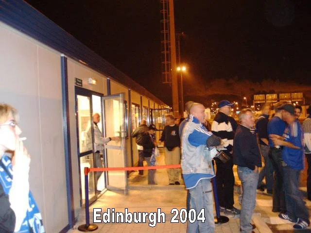 Menschen stehen in einer Warteschlange vor einem Gebäude bei Nacht, Schilder und Straßenlaternen sind sichtbar. Im Bild sind Männer und Frauen, einige tragen Kleidung im Blau- und Weißtönen. Text im Bild: "Edinburgh 2004".