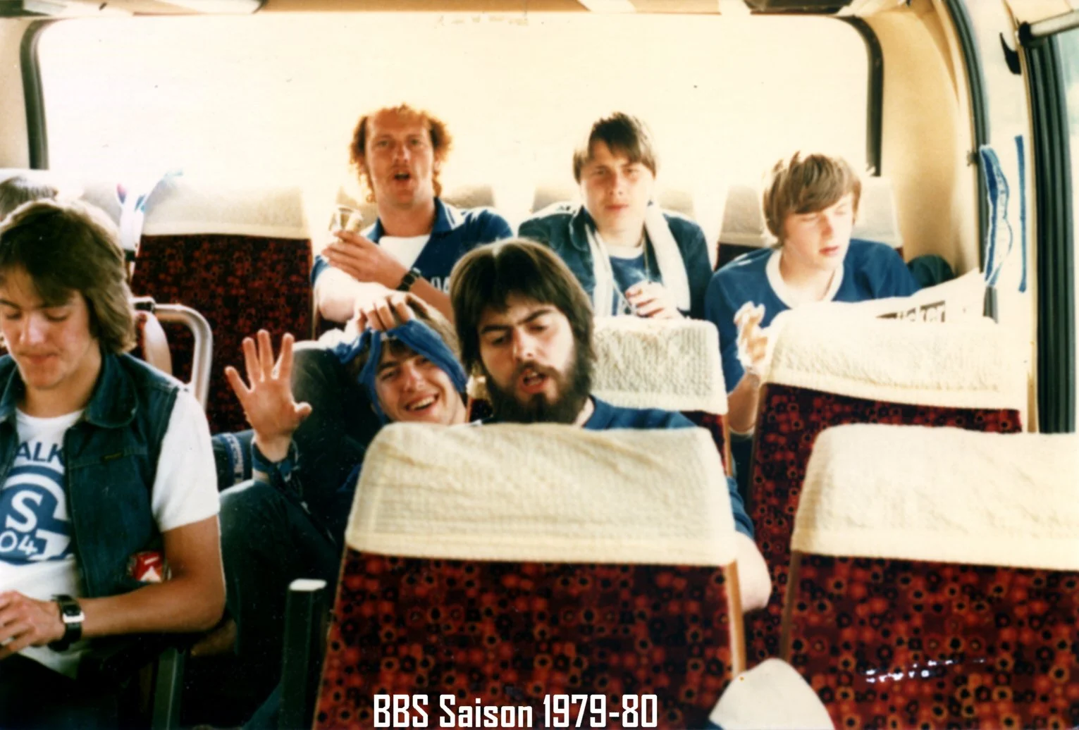 Jugendliche und junge Erwachsene sitzen in einem Bus, einige lachen und machen Grimassen, sie tragen Freizeitkleidung wie T-Shirts und Jacken. Der Text am unteren Rand lautet 'BBS Saison 1979-80'.