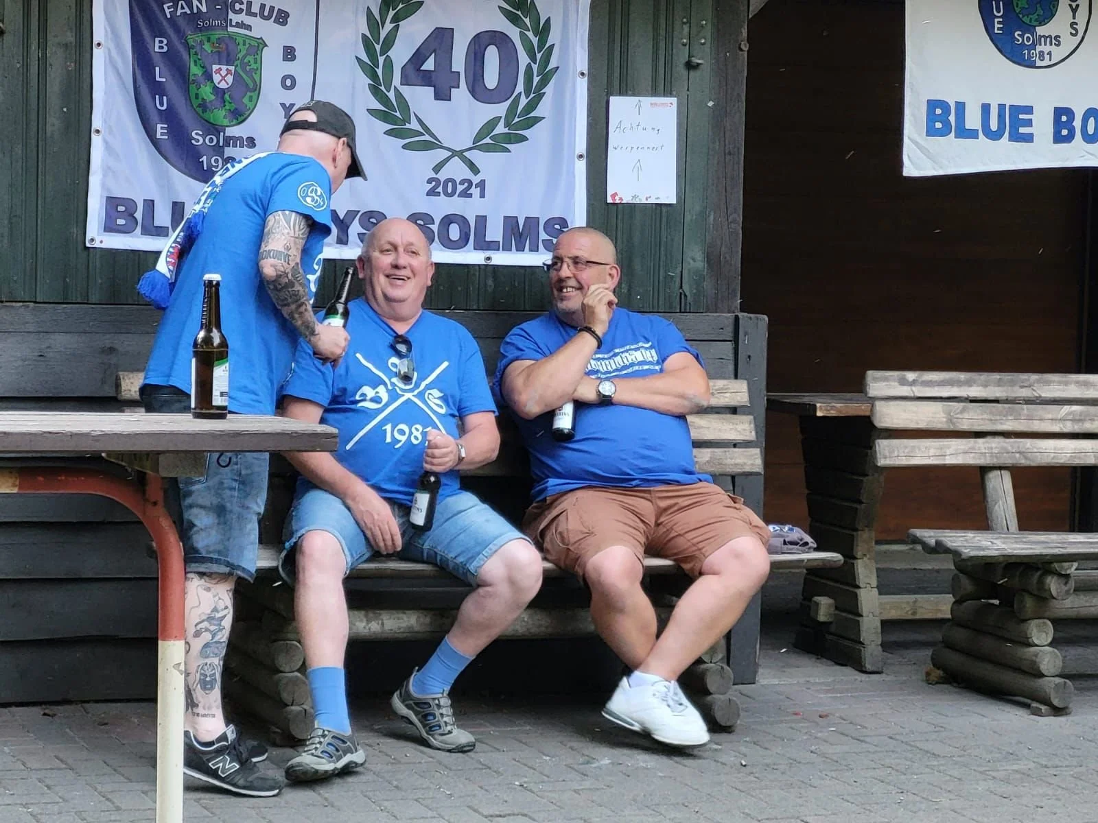 Drei Männer sitzen auf einer Bank, feiern eine Vereinsgründung oder Jubiläum, tragen blaue Shirts mit einem Logo, mit Bierflaschen in der Hand, es hängt eine Flagge mit der Aufschrift '40' und '2021' im Hintergrund.