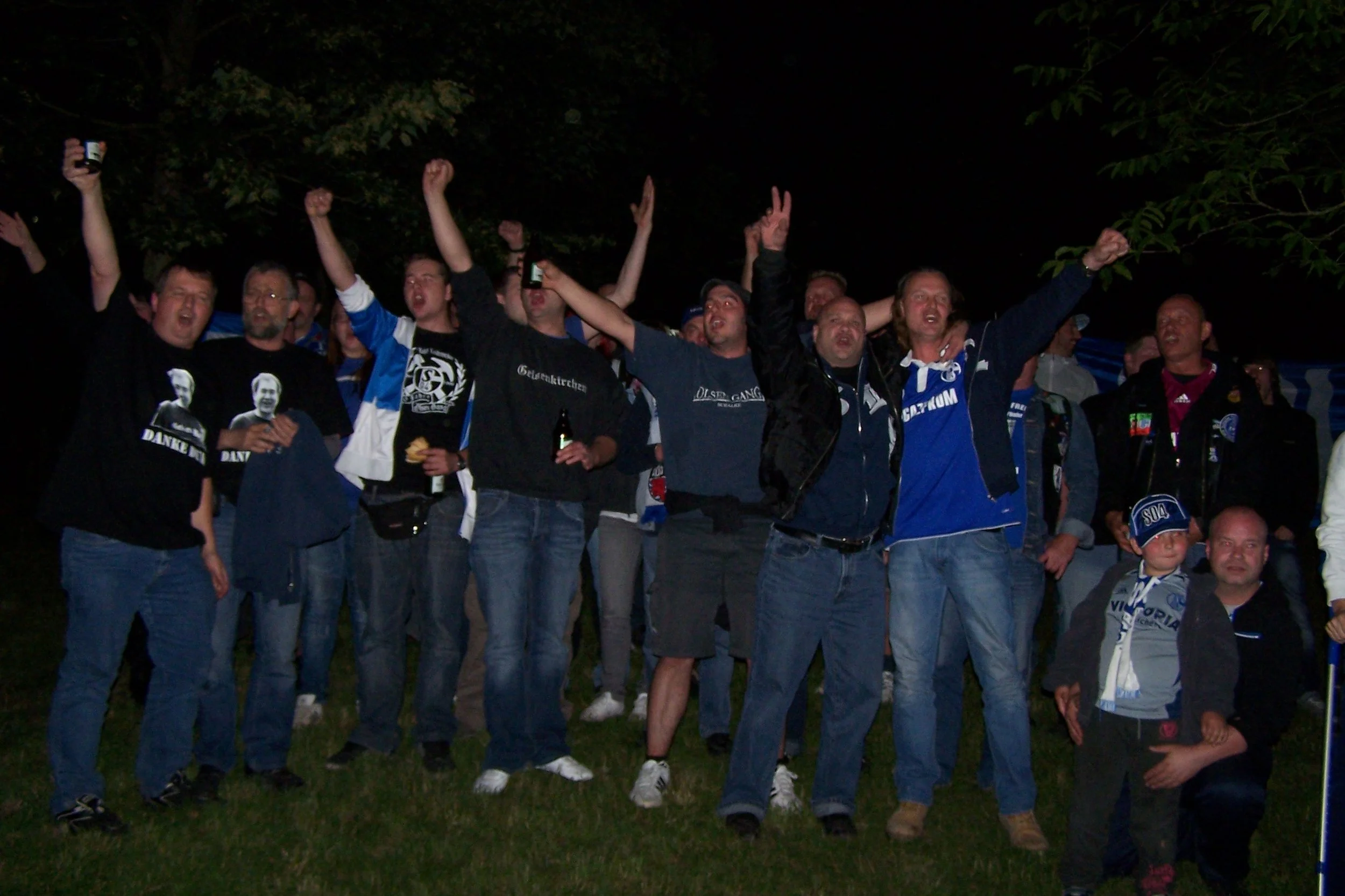 Gruppe von Menschen, die draußen in der Nacht feiern oder jubeln, einige tragen dunkle Kleidung mit Aufdrucken, einige halten Getränke in der Hand, einige machen Peace-Zeichen, im Hintergrund sind dunkle Bäume.