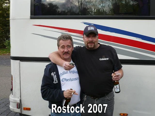 Zwei Männer stehen nebeneinander vor einem Bus, umarmen sich und lachen. Der Mann auf der linken trägt eine Jacke und ein T-Shirt mit blauem Text, der Mann auf der rechten trägt ein schwarzes T-Shirt mit Guinness-Logo und eine dunkle Kappe mit der Za